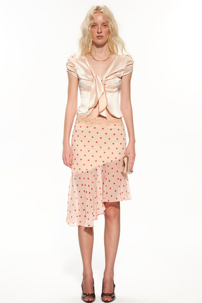 A model stands against a white studio background wearing a light peach satin top with a large front bow and a matching light peach polka-dot skirt with a lace overlay.
