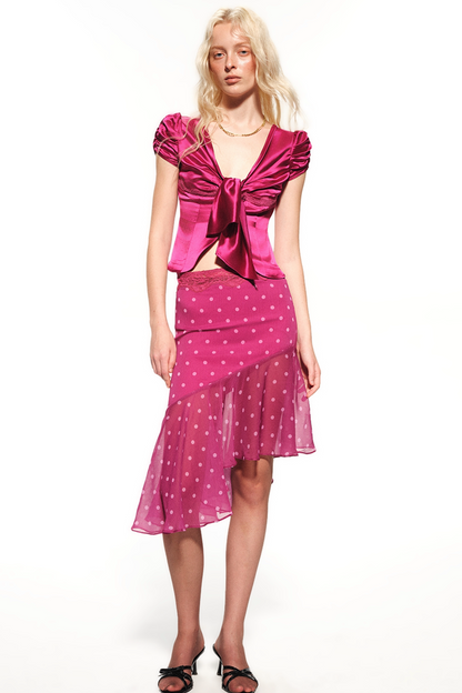 A blonde model wearing a vibrant magenta pink two-piece set consisting of a ruched short-sleeve top with a large front bow and a matching polka-dot skirt, paired with black strappy heels against a plain white background.