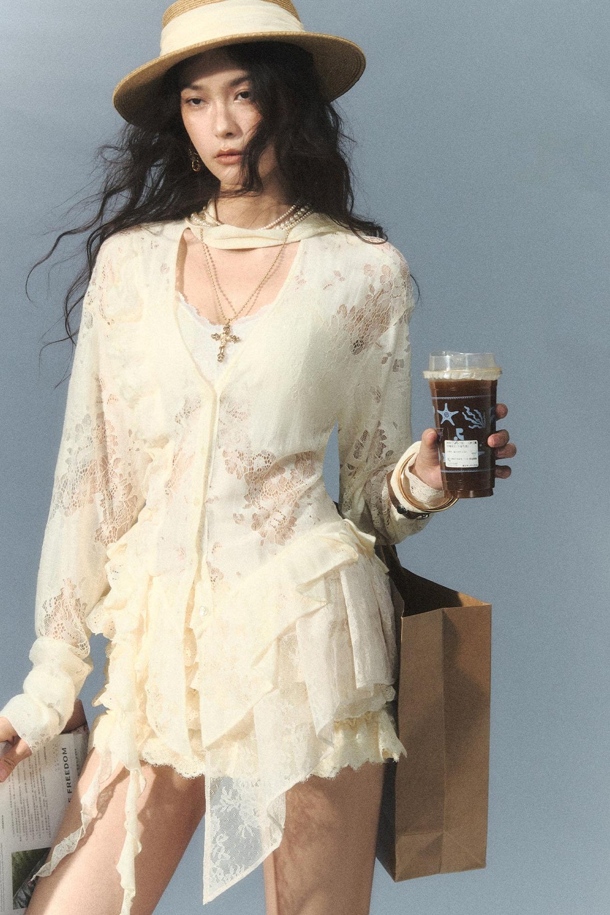 A woman wearing a cream lace long-sleeve top and matching hat, holding an iced coffee and a shopping bag against a solid blue-gray background.