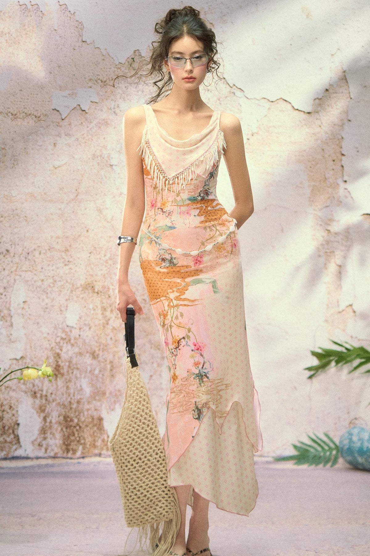 A woman models a sleeveless, asymmetrical maxi dress with a beige base and orange floral print, holding a woven straw bag against a textured, rustic wall.
