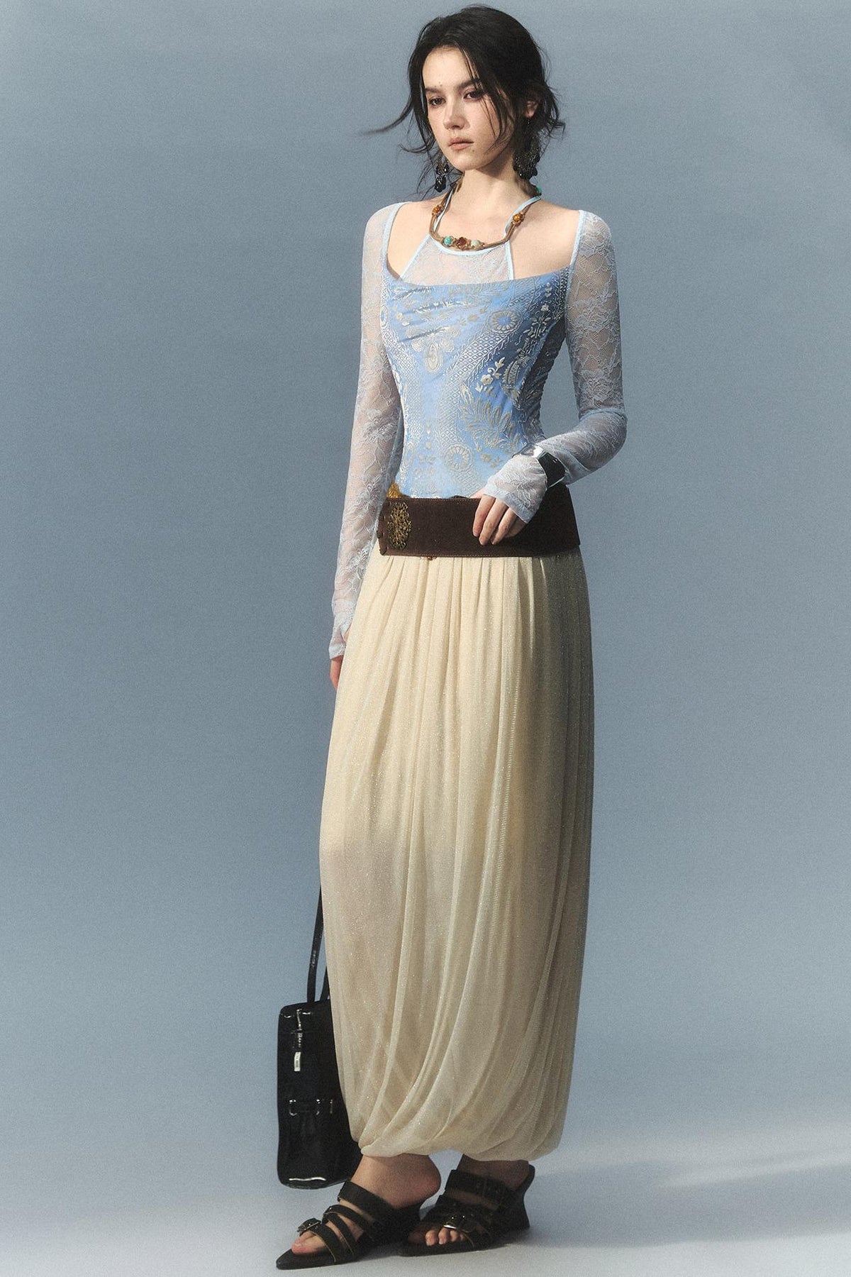 A model stands against a light blue-gray studio background wearing a long-sleeve lace-trimmed top with a light blue embroidered bodice, a long cream pleated skirt, a dark brown belt, and black sandals, while holding a small black handbag.