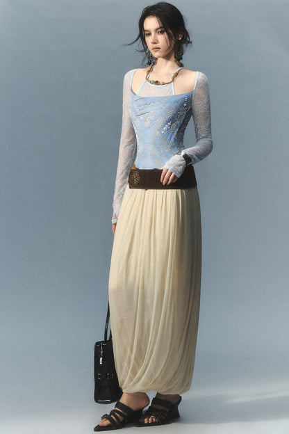 A model stands against a light blue-gray studio background wearing a long-sleeve lace-trimmed top with a light blue embroidered bodice, a long cream pleated skirt, a dark brown belt, and black sandals, while holding a small black handbag.