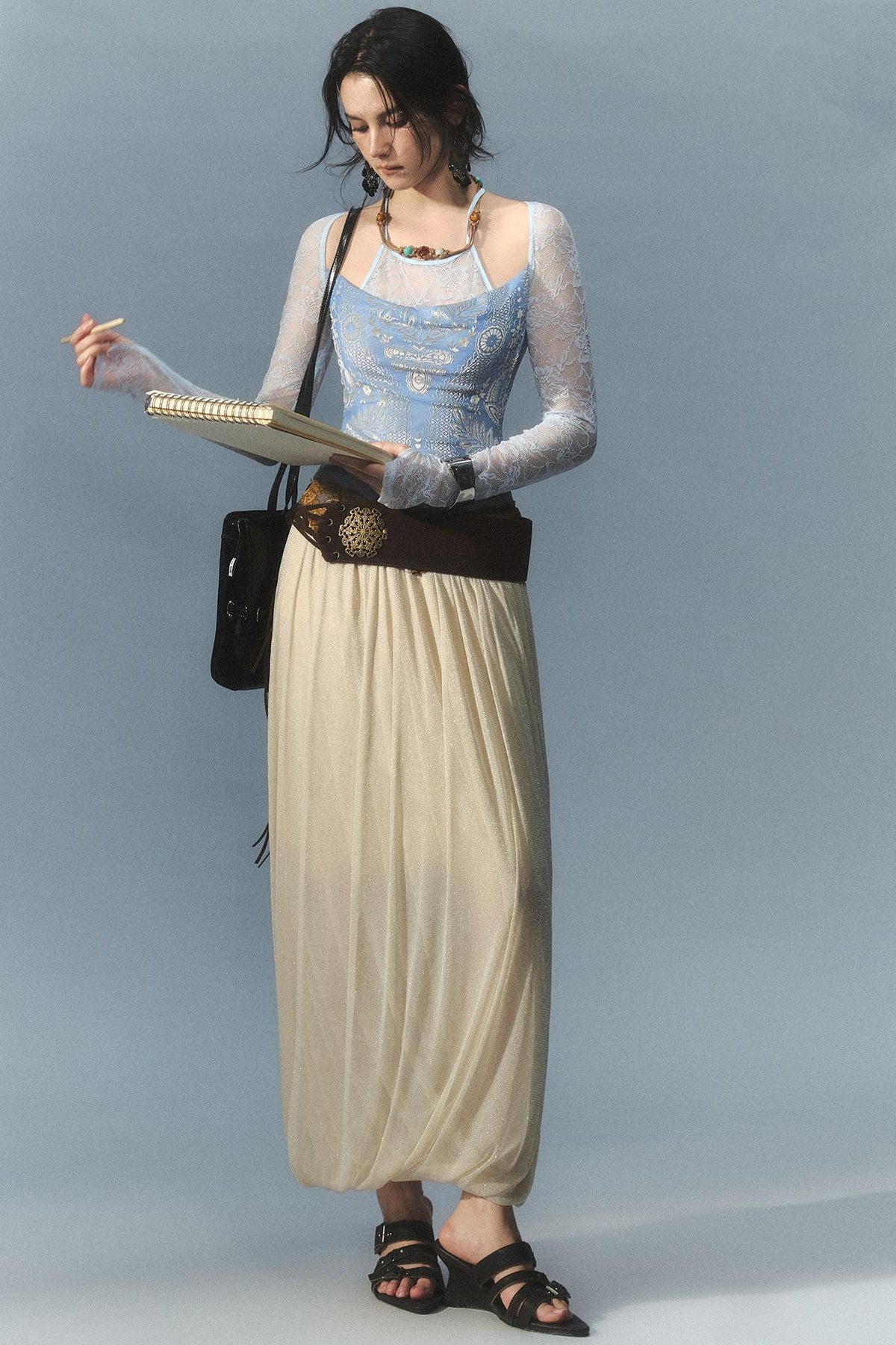 A woman stands against a light blue-gray studio background wearing a long-sleeve cream maxi skirt with a wide dark brown belt, a light blue embroidered camisole, sheer white long sleeves, white gloves, and black sandals. She holds a small notebook or book and carries a black shoulder bag.