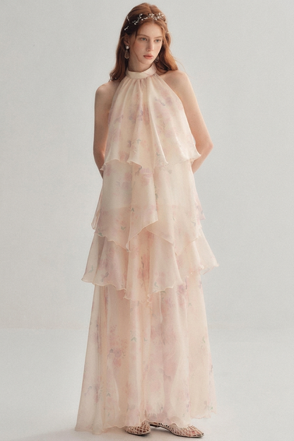 A woman models a sleeveless, high-neck, tiered maxi dress in a soft blush pink color with a delicate floral print. The dress features multiple ruffled layers and is paired with a matching floral headband and flat sandals, set against a plain light gray studio background.