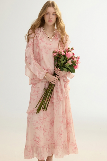 A woman modeling a long, flowing light pink dress with a soft floral print, holding a bouquet of pink flowers against a plain light background.