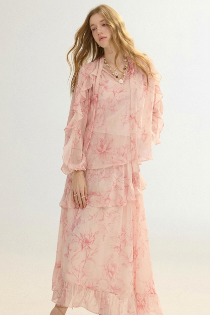 A woman modeling a light pink and white floral print two-piece set consisting of a long-sleeve ruffled blouse and a matching tiered maxi skirt, set against a plain light background.