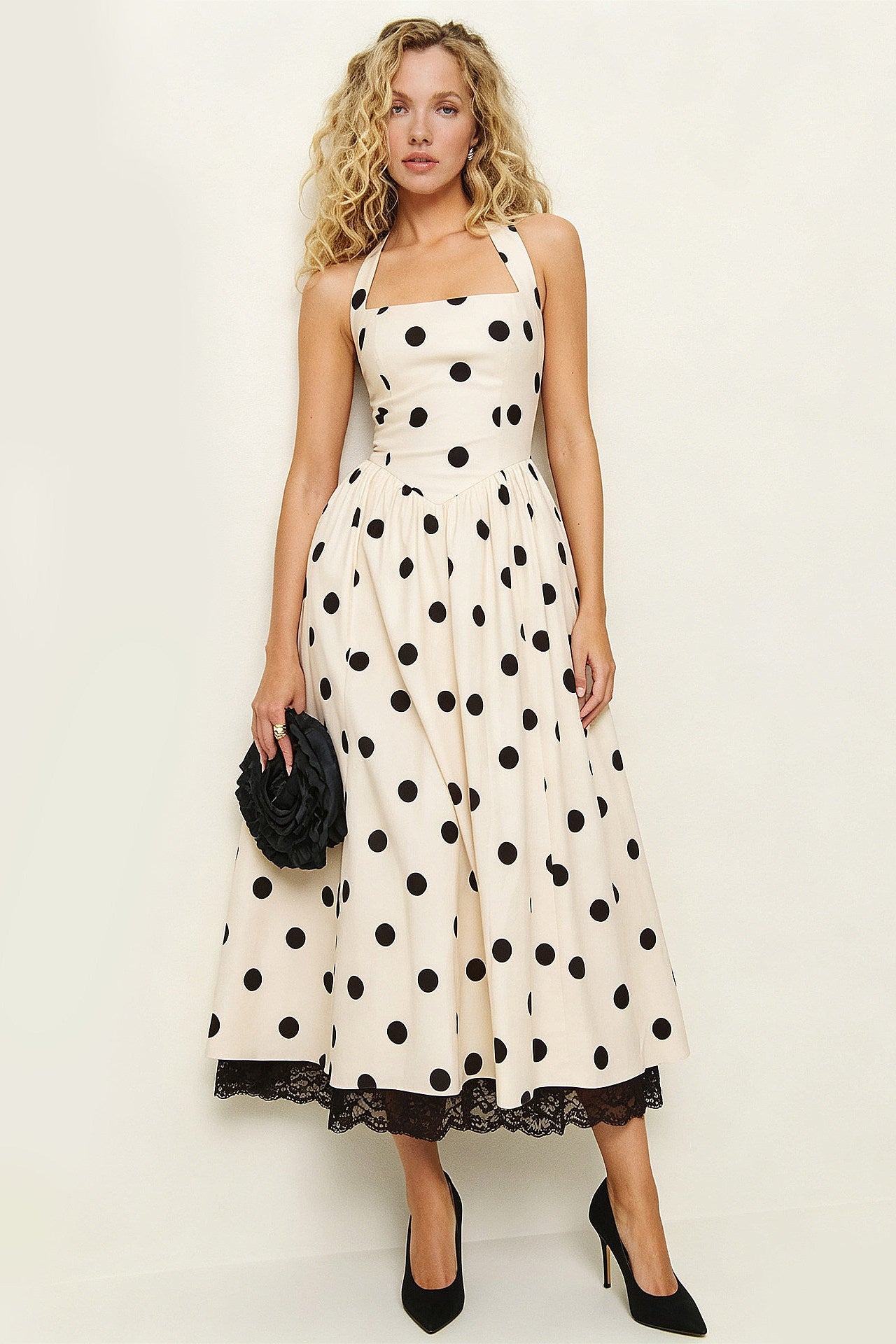 A woman modeling a sleeveless cream midi dress with black polka dots, a square neckline, a fitted bodice, and a flared skirt. The dress has black lace trim at the hem and is styled with black heels and a small black clutch against a plain off-white studio background.