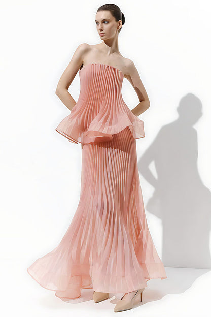 A model wears a strapless pleated gown in a soft peach-pink color, standing against a white studio background with a prominent shadow cast to the right.