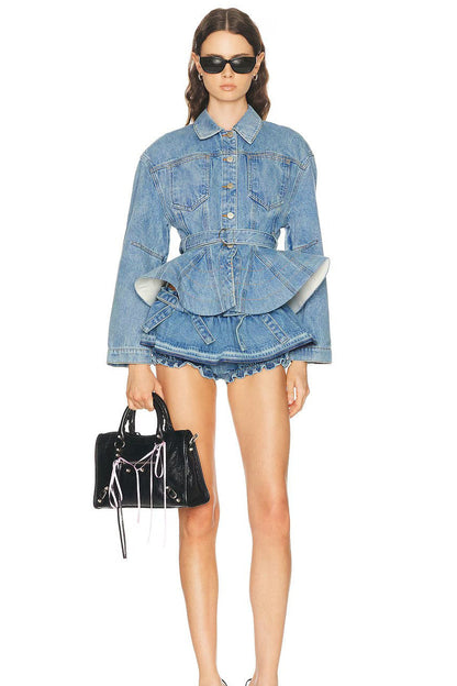 A model wearing a light blue denim jacket with a unique layered peplum waist and matching frayed denim shorts, styled with black sunglasses and a black handbag with silver chains against a plain white background.