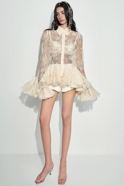 A model wearing a short, sheer lace blouse with long sleeves, a high collar, and a ruffled tulle hem in a light cream color, paired with clear high-heeled sandals against a plain white studio background.
