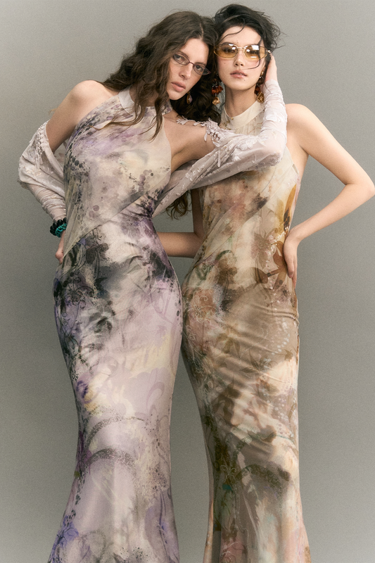 Two women modeling floor-length sheer satin dresses with abstract painterly prints in soft lavender, beige, gray, and brown tones against a plain gray studio background.
