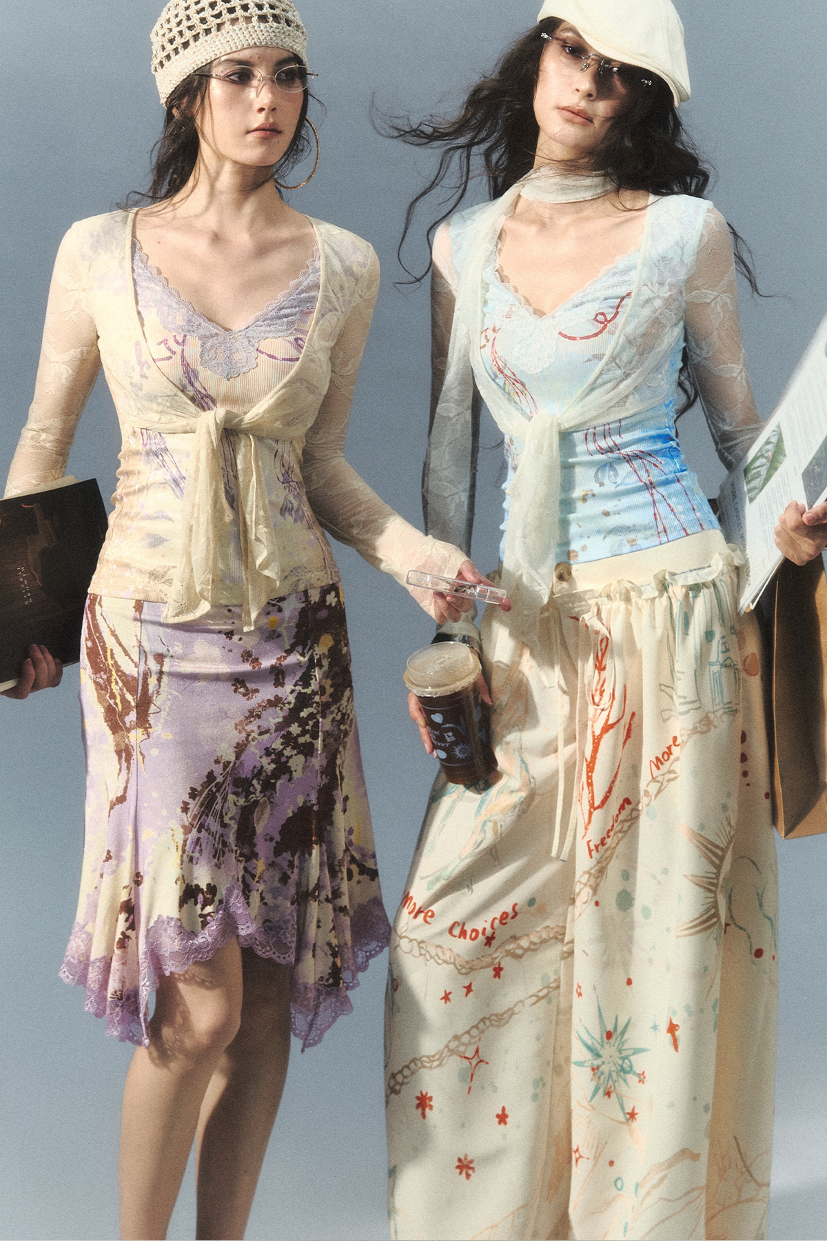Two women modeling coordinated printed outfits against a plain light gray studio background. The woman on the left wears a cream top with a purple floral skirt and a crocheted hat, while the woman on the right wears a pale blue top with a cream skirt featuring orange and blue botanical prints.