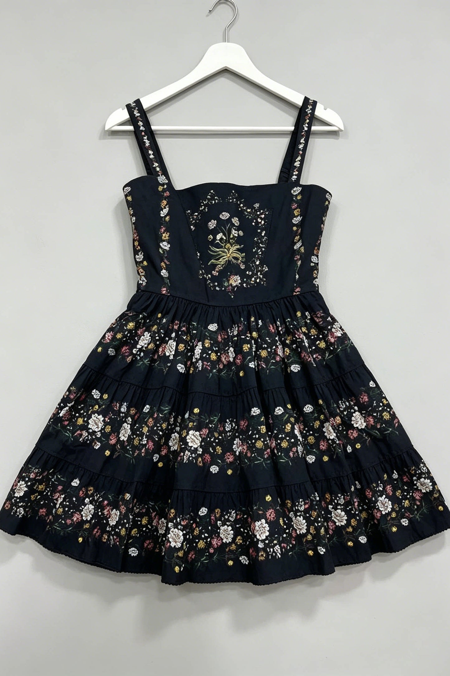A sleeveless black dress with floral embroidery and applique, hanging on a white plastic hanger against a plain light gray wall.