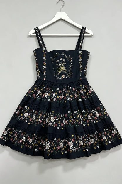 A sleeveless black dress with floral embroidery and applique, hanging on a white plastic hanger against a plain light gray wall.