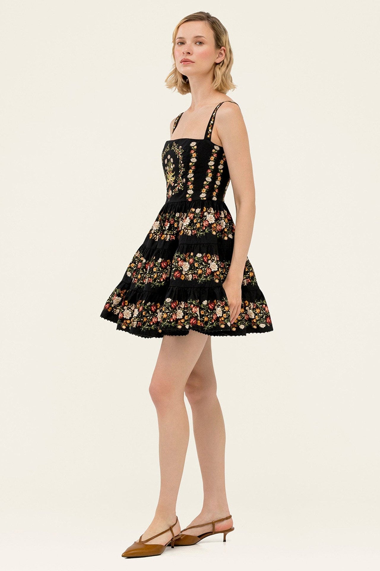 A woman modeling a short black dress with floral embroidery and thin shoulder straps, paired with brown slingback heels, against a plain light beige studio background.