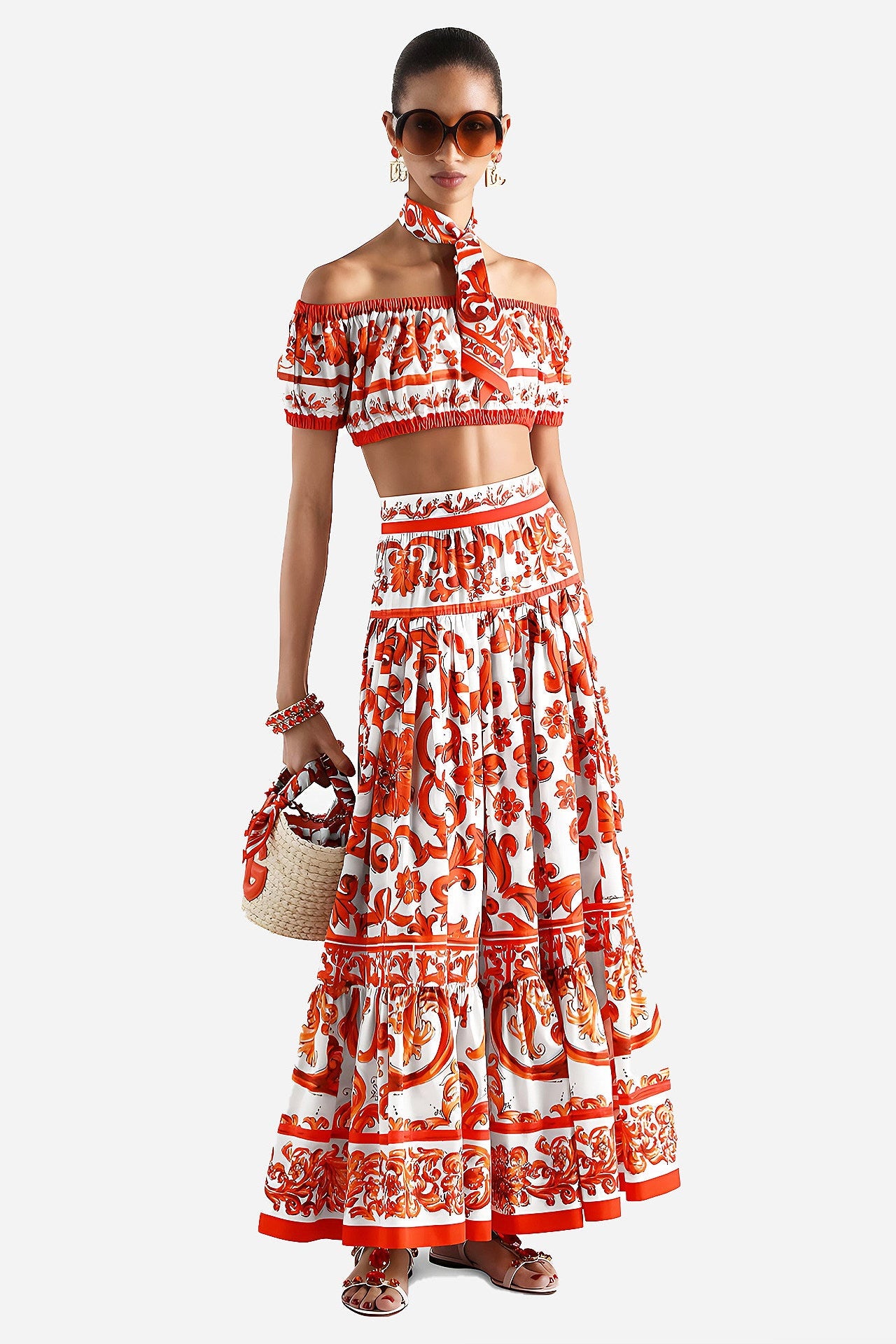 A model wearing a matching off-the-shoulder crop top and tiered maxi skirt set in a white fabric with a bold red and orange floral print, styled with sunglasses, a woven straw bag, and sandals against a light gray background.