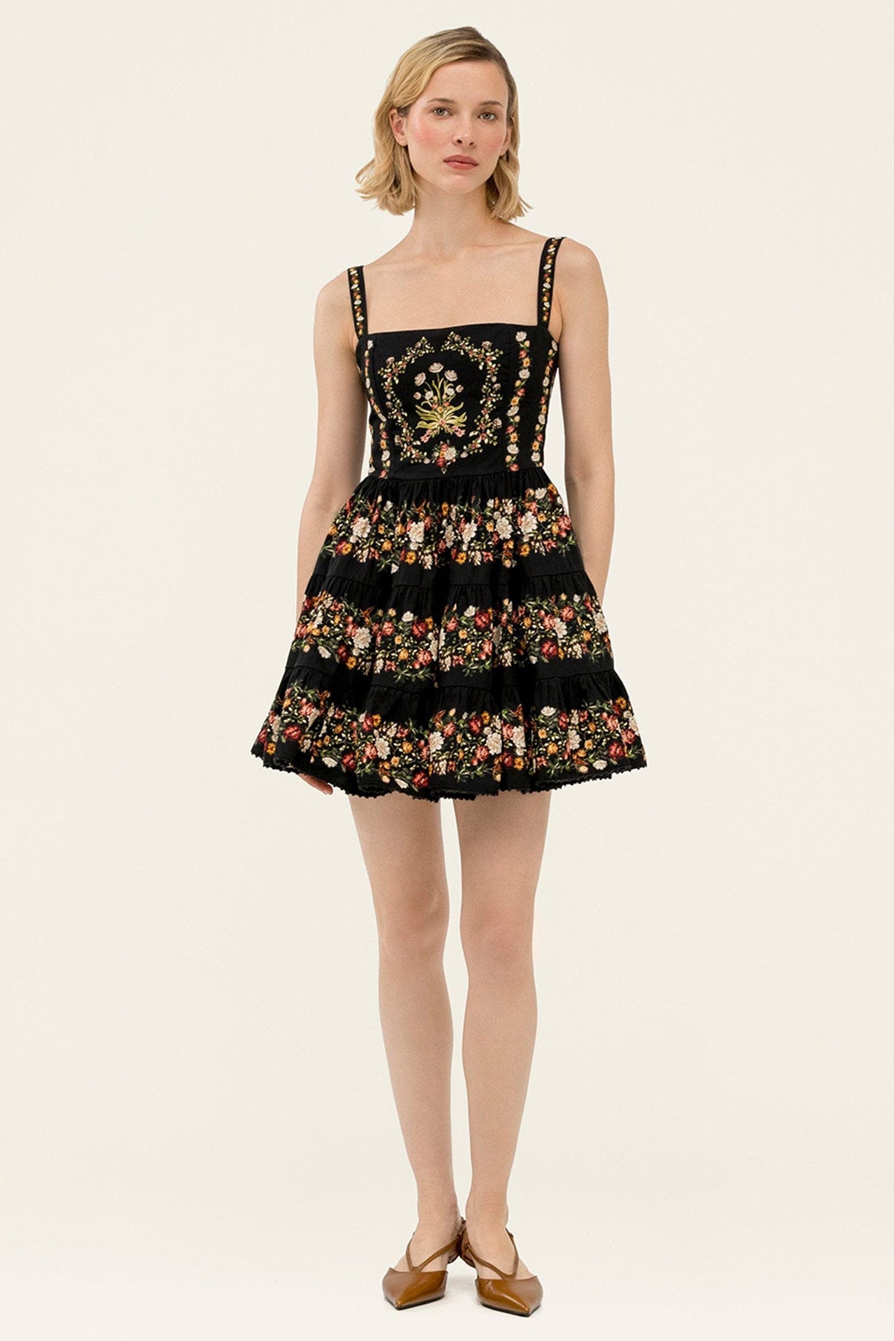 A woman modeling a short black dress with floral embroidery and gold embellishments on the bodice, photographed against a plain light beige studio background.