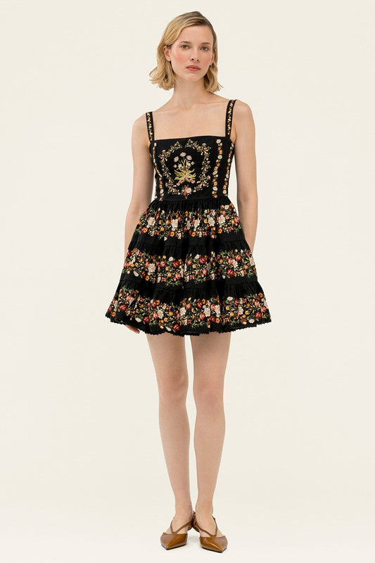 A woman modeling a short black dress with floral embroidery and gold embellishments on the bodice, photographed against a plain light beige studio background.