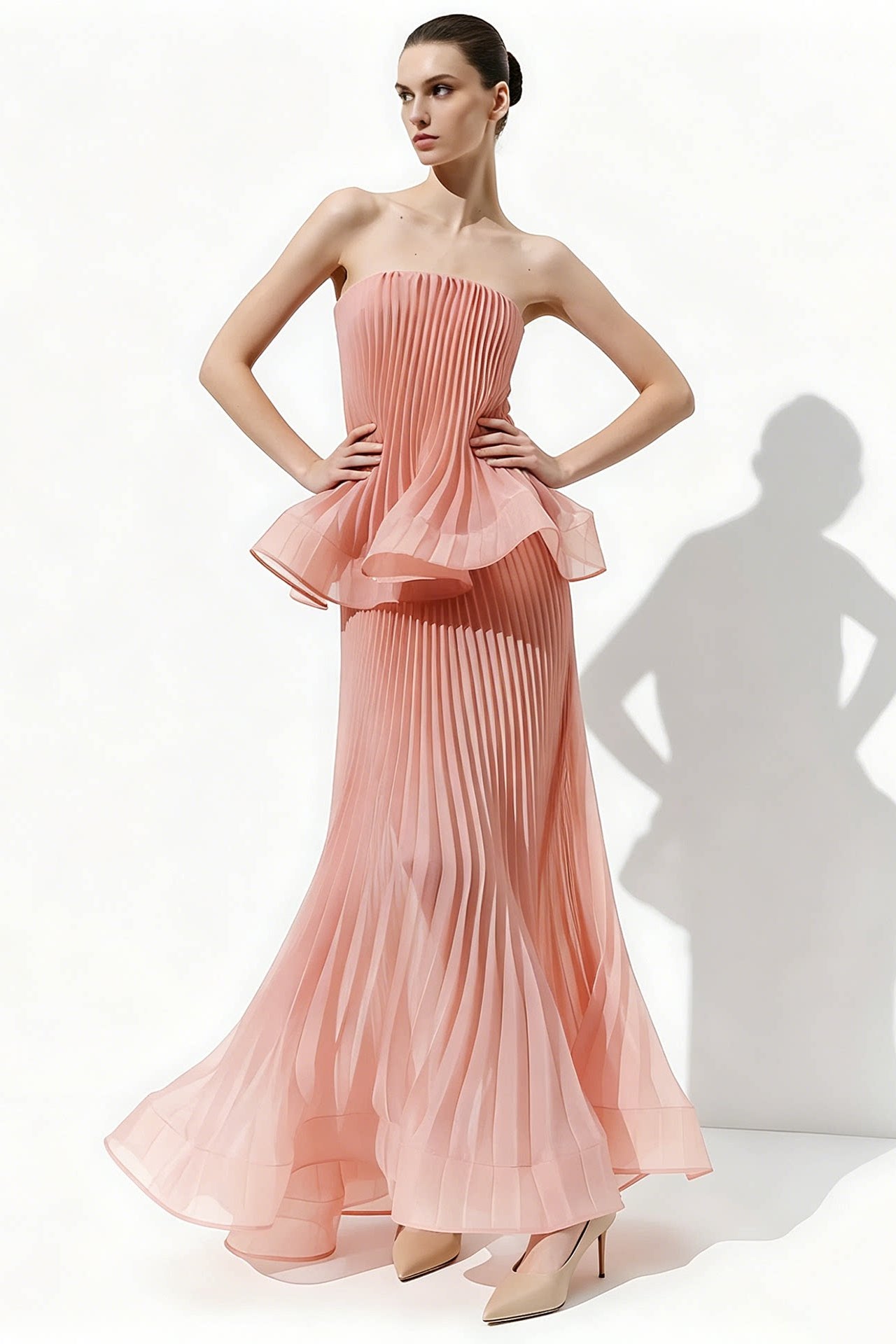 A woman models a strapless pleated maxi dress in a soft peach-pink color, standing against a white studio background with a prominent shadow cast to the right.