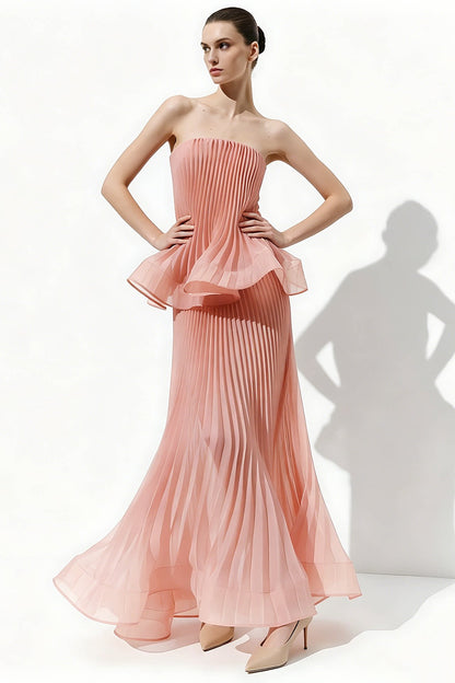 A woman models a strapless pleated maxi dress in a soft peach-pink color, standing against a white studio background with a prominent shadow cast to the right.