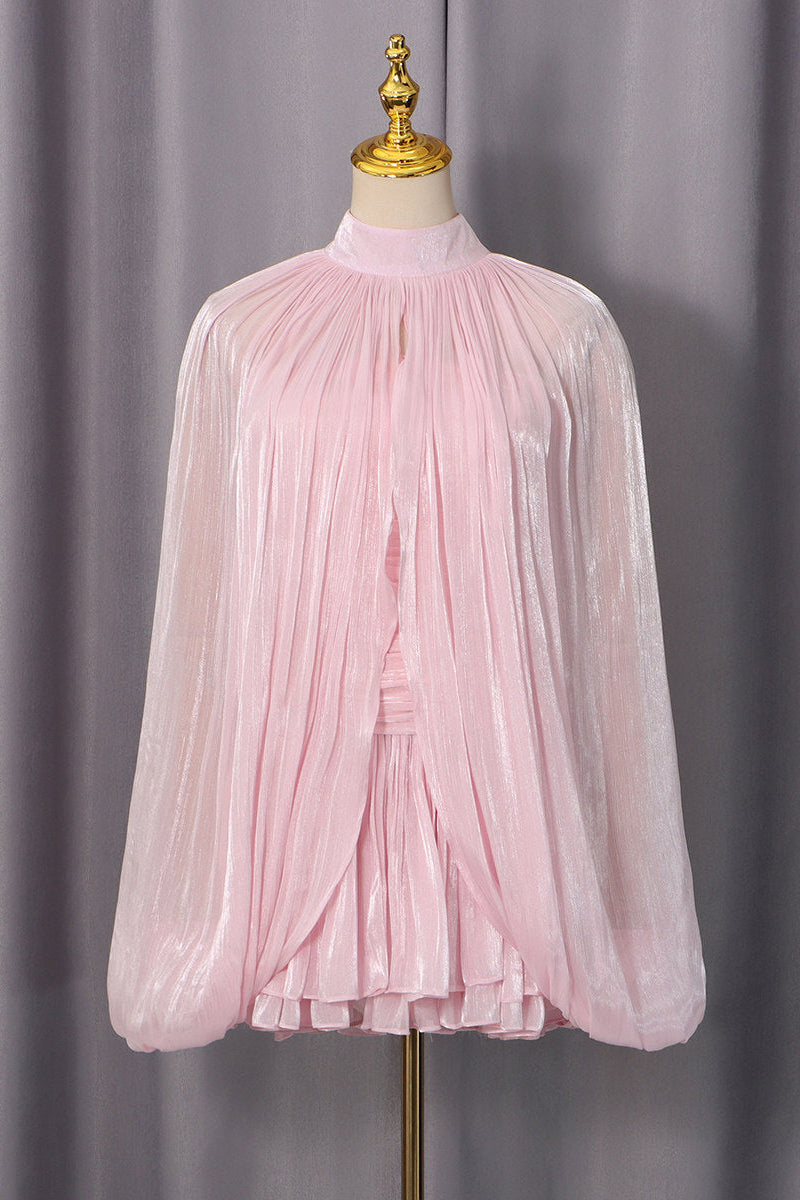 A light pink long-sleeve blouse with a high mock neck and dramatic pleated, crinkled fabric, displayed on a cream dress form with a gold finial against a gray curtain backdrop.