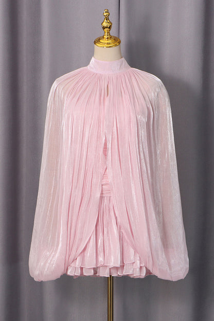 A light pink long-sleeve blouse with a high mock neck and dramatic pleated, crinkled fabric, displayed on a cream dress form with a gold finial against a gray curtain backdrop.