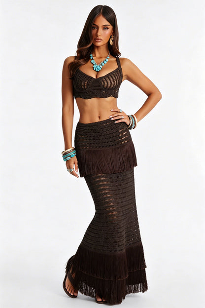 A woman models a dark brown crochet two-piece set consisting of a halter crop top and a fringed maxi skirt, accessorized with turquoise jewelry against a plain white background.