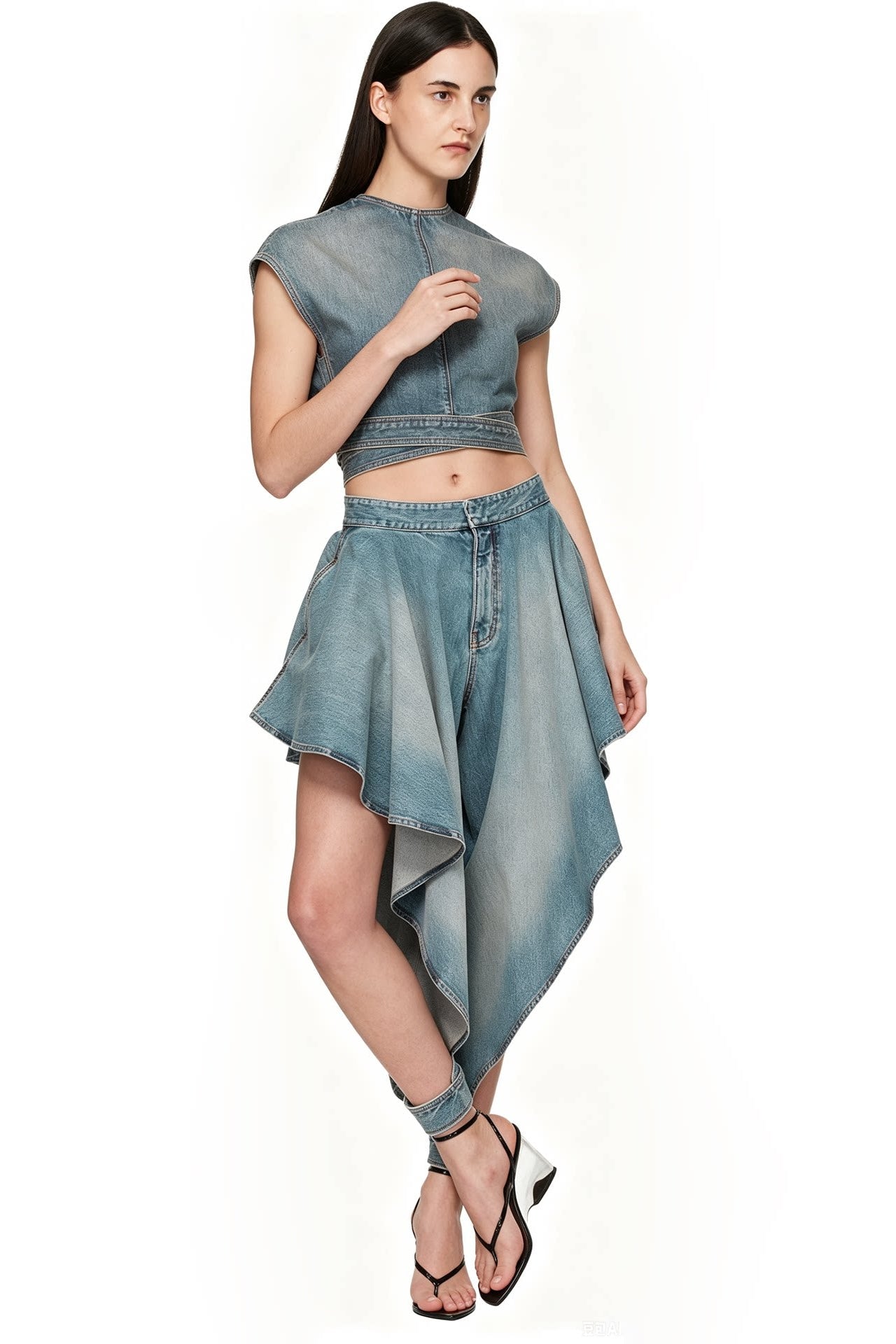 A model wearing a light blue denim two-piece set consisting of a cropped sleeveless top and an asymmetrical high-low skirt, paired with black strappy heels, against a plain white background.