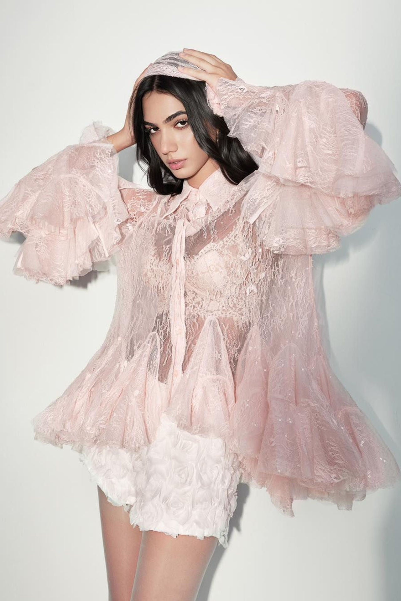 A model wearing a light pink sheer tulle jacket with dramatic ruffled sleeves, a matching headscarf, and a white floral lace mini skirt, posed against a plain light gray studio background.