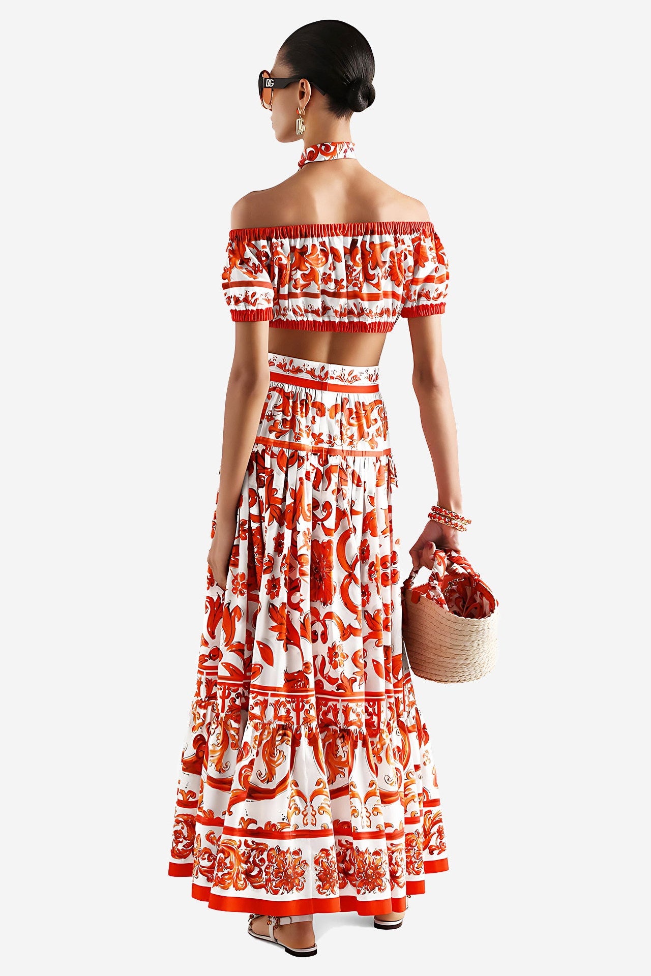 A woman shown from the back wearing a matching off-the-shoulder crop top and long skirt set in white with a bold red and orange floral pattern. She is holding a woven straw bag with a red fabric lining, set against a plain light gray studio background.