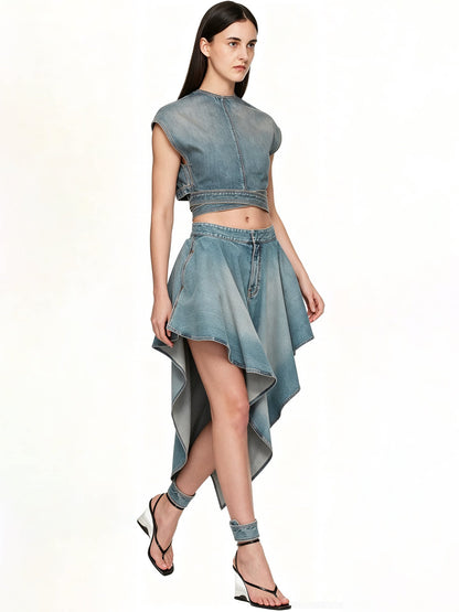 A model wearing a light blue denim two-piece set consisting of a cropped sleeveless top and a high-low skirt, paired with matching denim ankle cuffs and black strappy heels, against a plain white background.