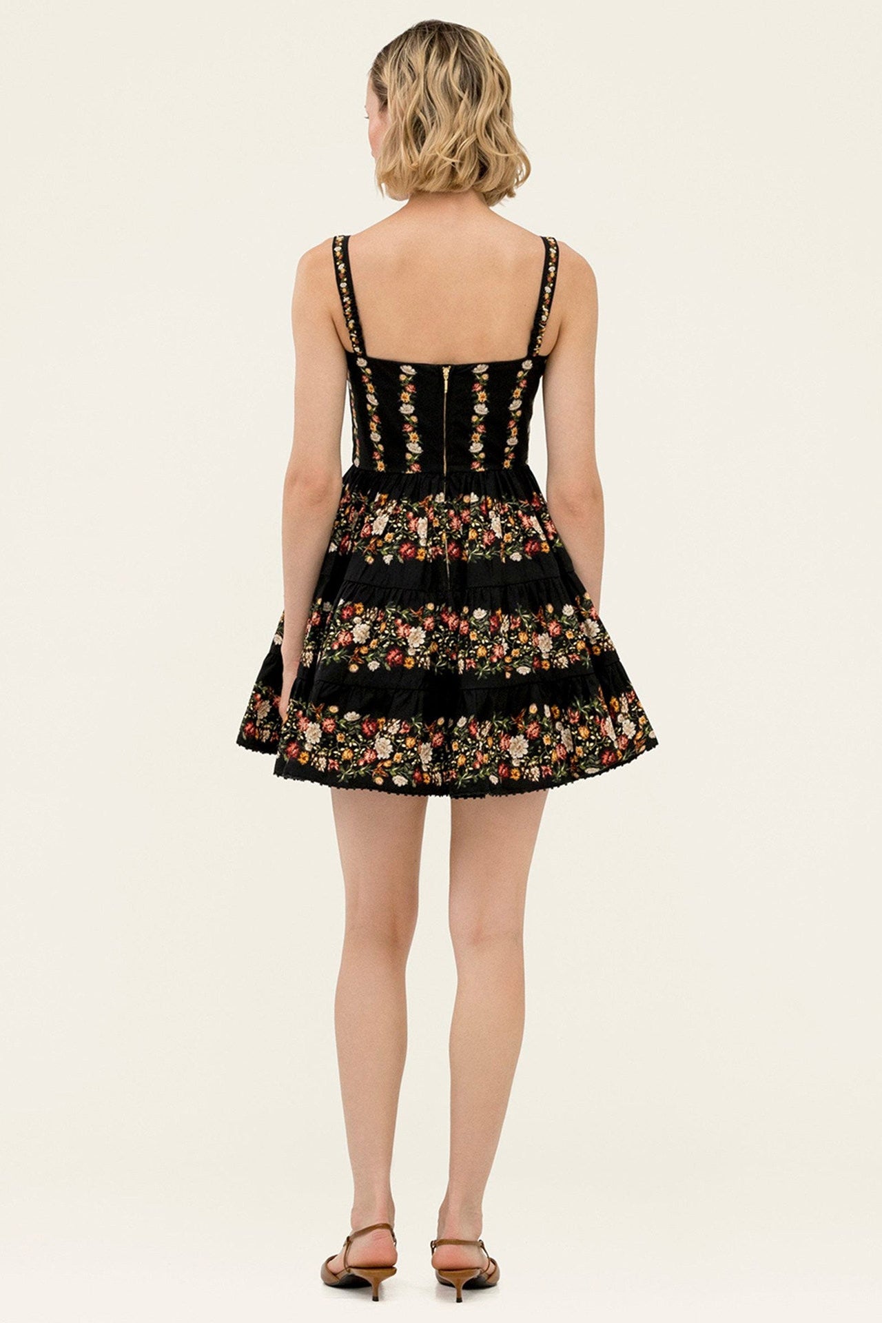 A woman viewed from the back wearing a sleeveless black dress with floral embroidery and thin shoulder straps, paired with brown heeled sandals, against a plain light beige studio background.