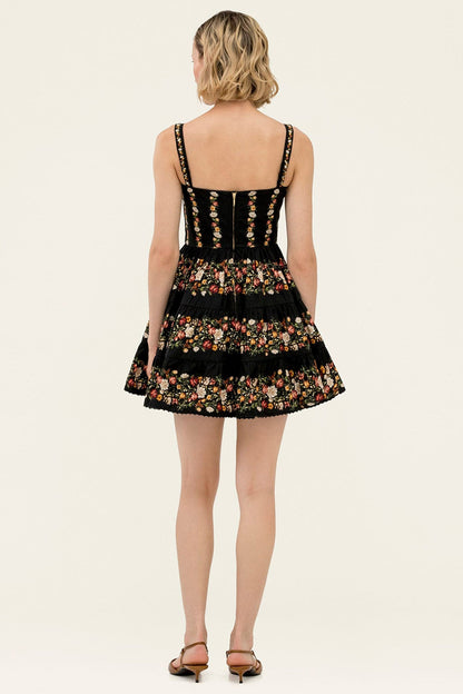 A woman viewed from the back wearing a sleeveless black dress with floral embroidery and thin shoulder straps, paired with brown heeled sandals, against a plain light beige studio background.