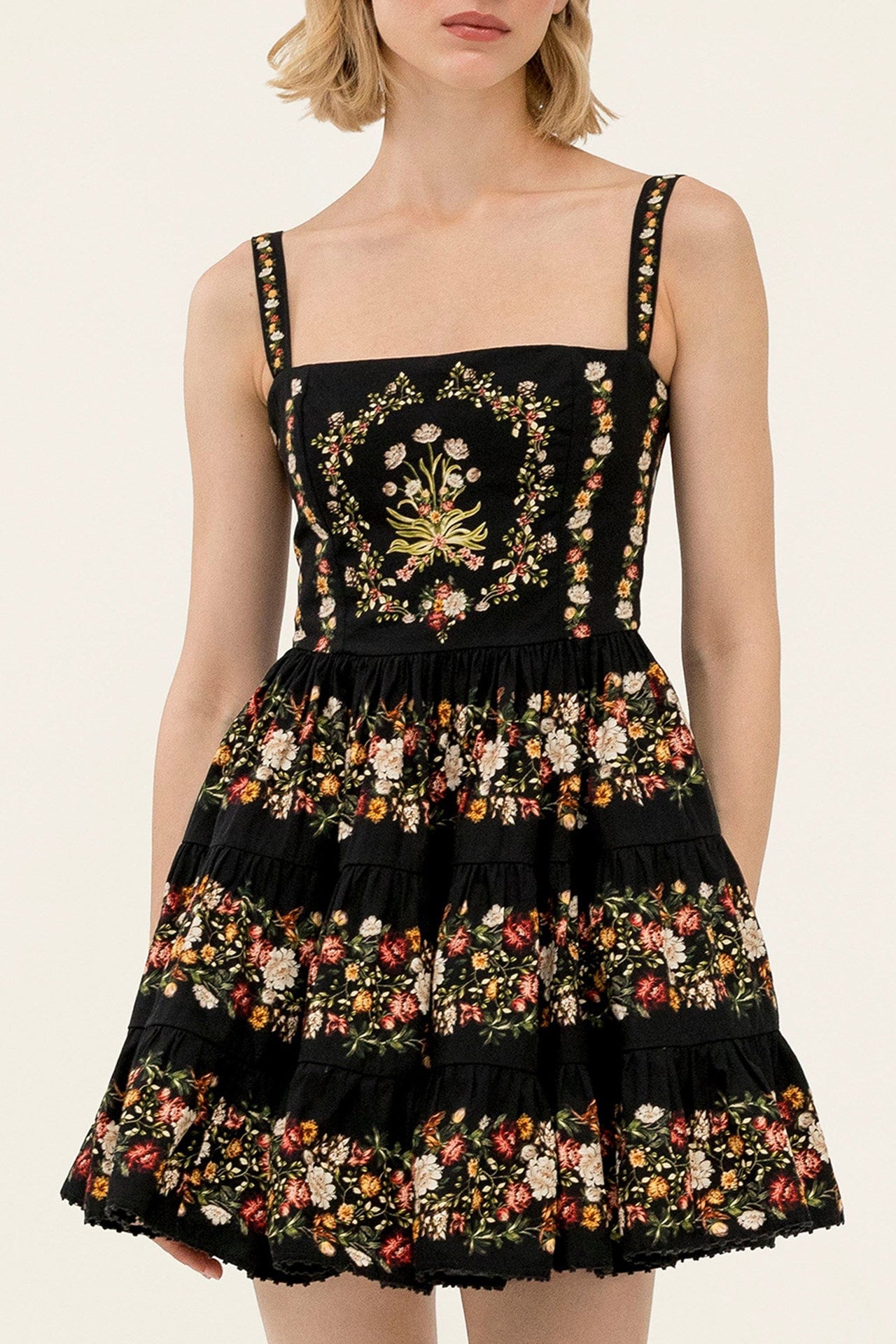 A model wearing a black tiered mini dress with a colorful floral print and an embroidered gold and green floral motif on the bodice, set against a plain light beige background.
