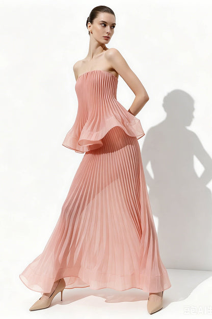 A woman models a strapless pleated maxi dress in a soft dusty pink color, standing against a white studio background with a prominent cast shadow.