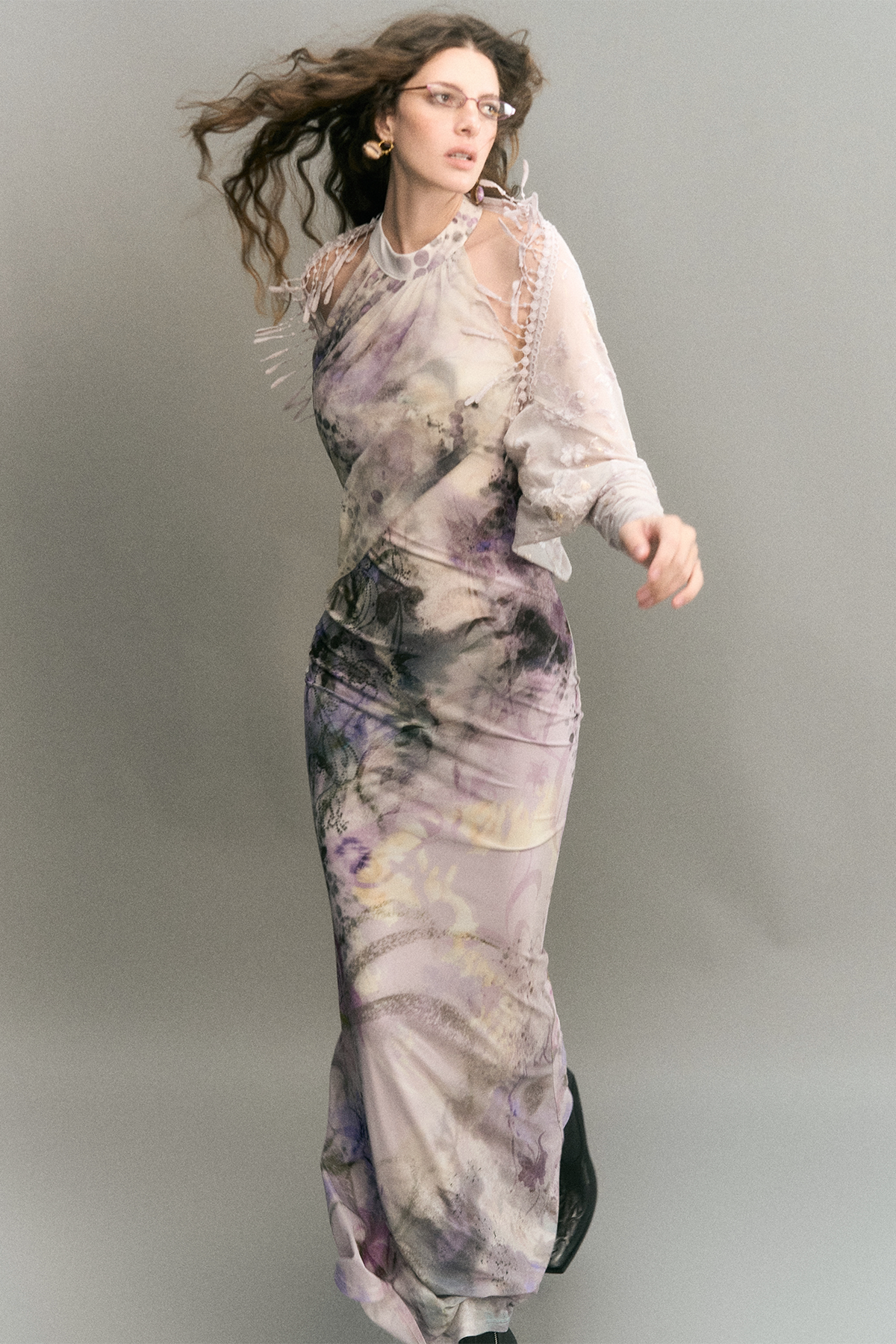 A woman modeling a sheer, long-sleeved maxi dress with an abstract watercolor-like print in lavender, purple, gray, and black, posed against a solid gray studio background.