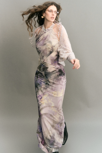 A woman modeling a sheer, long-sleeved maxi dress with an abstract watercolor-like print in lavender, purple, gray, and black, posed against a solid gray studio background.