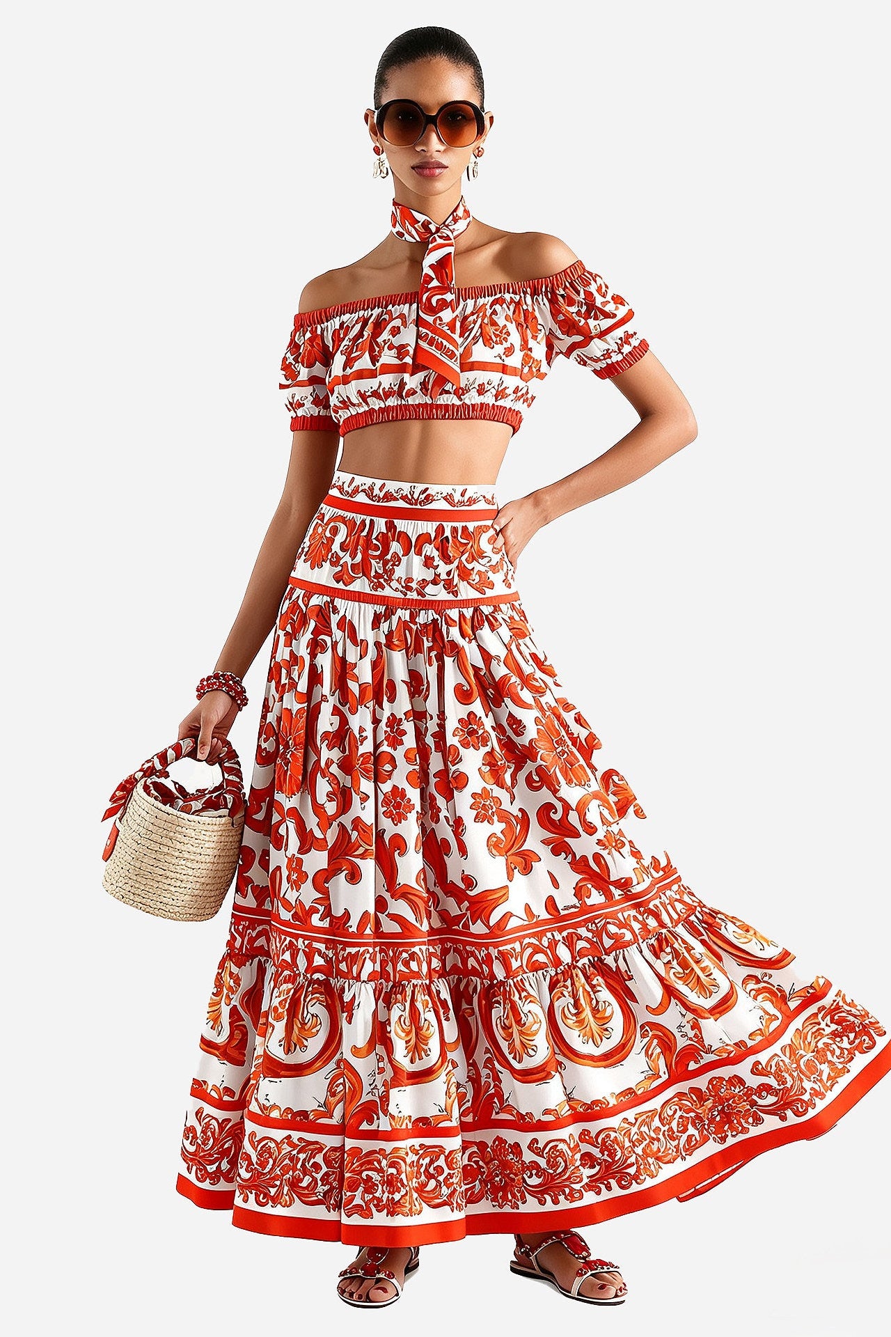 A woman modeling a matching off-the-shoulder crop top and long tiered maxi skirt set in a red and white paisley print, holding a woven straw bag against a light gray studio background.