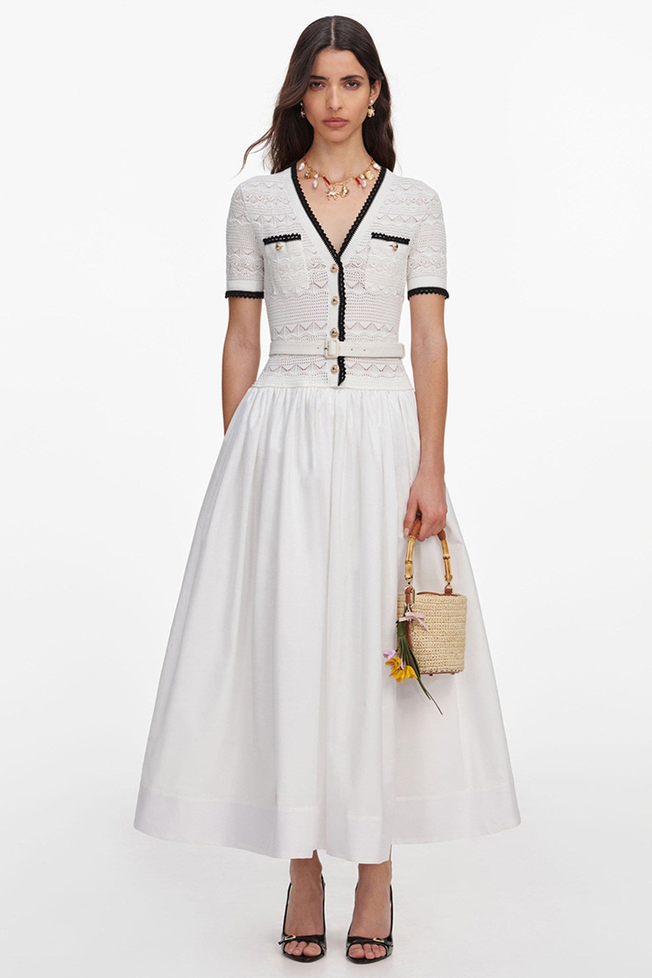 A model wears a white short-sleeve midi dress with black trim and lace detailing on the bodice, paired with a full white skirt. She is holding a small woven straw bucket bag and wearing black strappy heels against a plain white studio background.