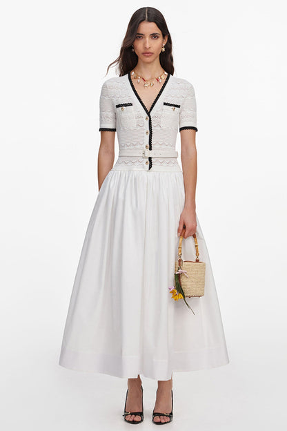 A model wears a white short-sleeve midi dress with black trim and lace detailing on the bodice, paired with a full white skirt. She is holding a small woven straw bucket bag and wearing black strappy heels against a plain white studio background.