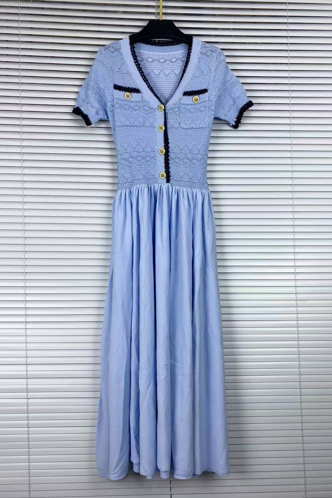 A light blue midi dress with a lace bodice, short sleeves, and gold buttons, hanging on a black hanger against a white window blind background.