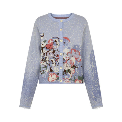 A light blue cropped cardigan sweater featuring a large multicolor floral tapestry design on the front, with gold buttons and a ribbed crew neck, photographed on a solid white background.