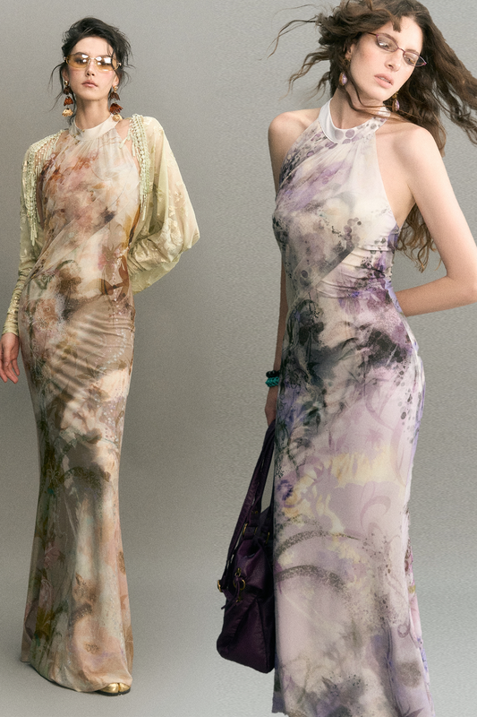 Two models wearing long, floor-length abstract-print gowns against a plain gray studio background. The dress on the left has a warm beige, tan, and brown watercolor-like pattern with cream sleeves.