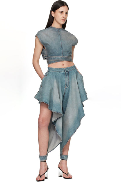 A model wearing a light blue denim two-piece outfit consisting of a cropped, asymmetrical cap-sleeve top and a matching high-low skirt, paired with black strappy heels against a white background.