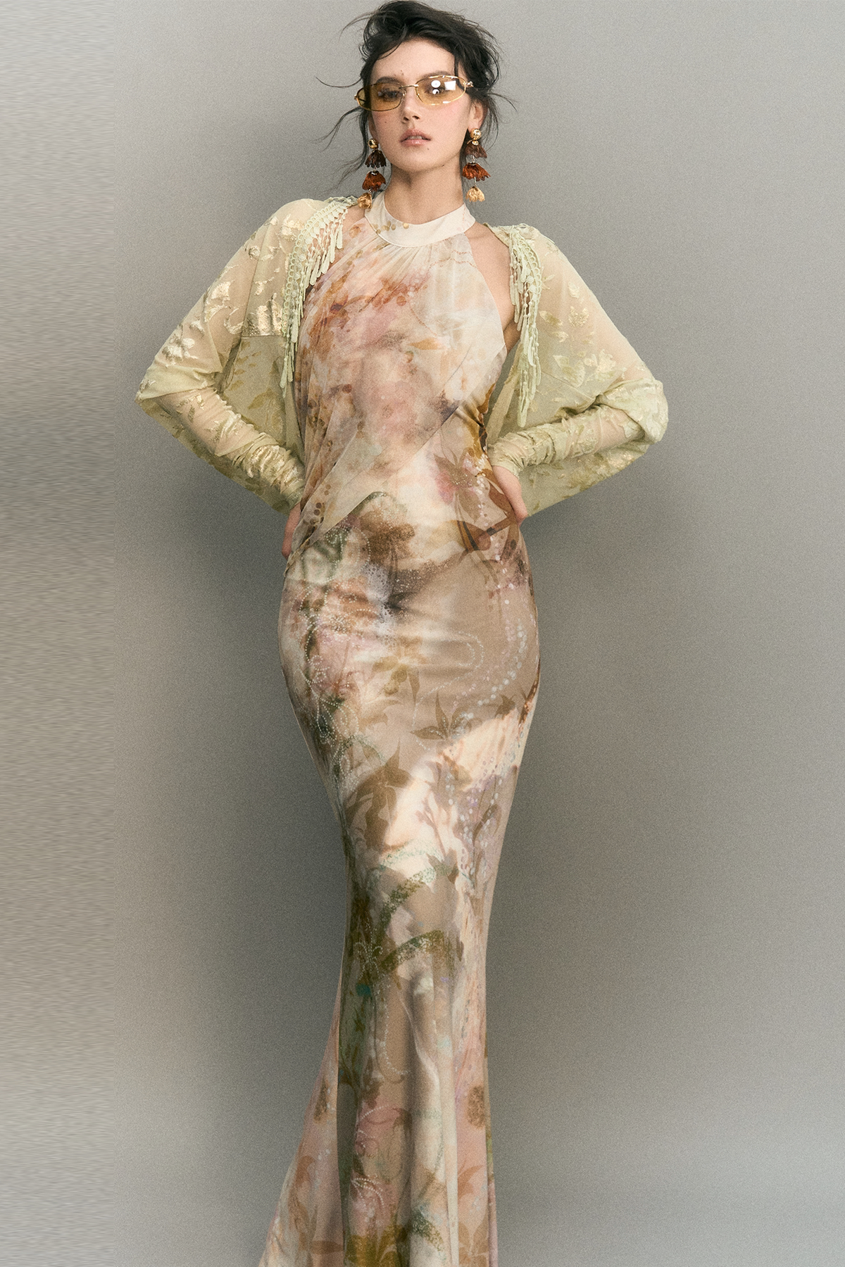 A model wearing a floor-length beige and brown floral print gown with a matching sheer long-sleeve shrug and gold-framed sunglasses, posing against a neutral gray studio background.