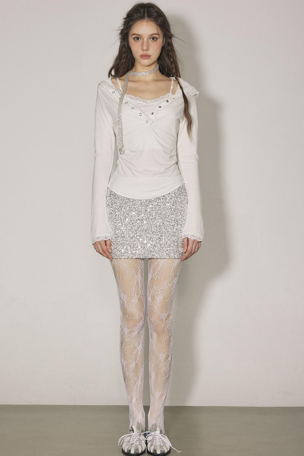 Woman wearing a white long-sleeve top, silver sequin skirt, and sheer stockings against a plain background
