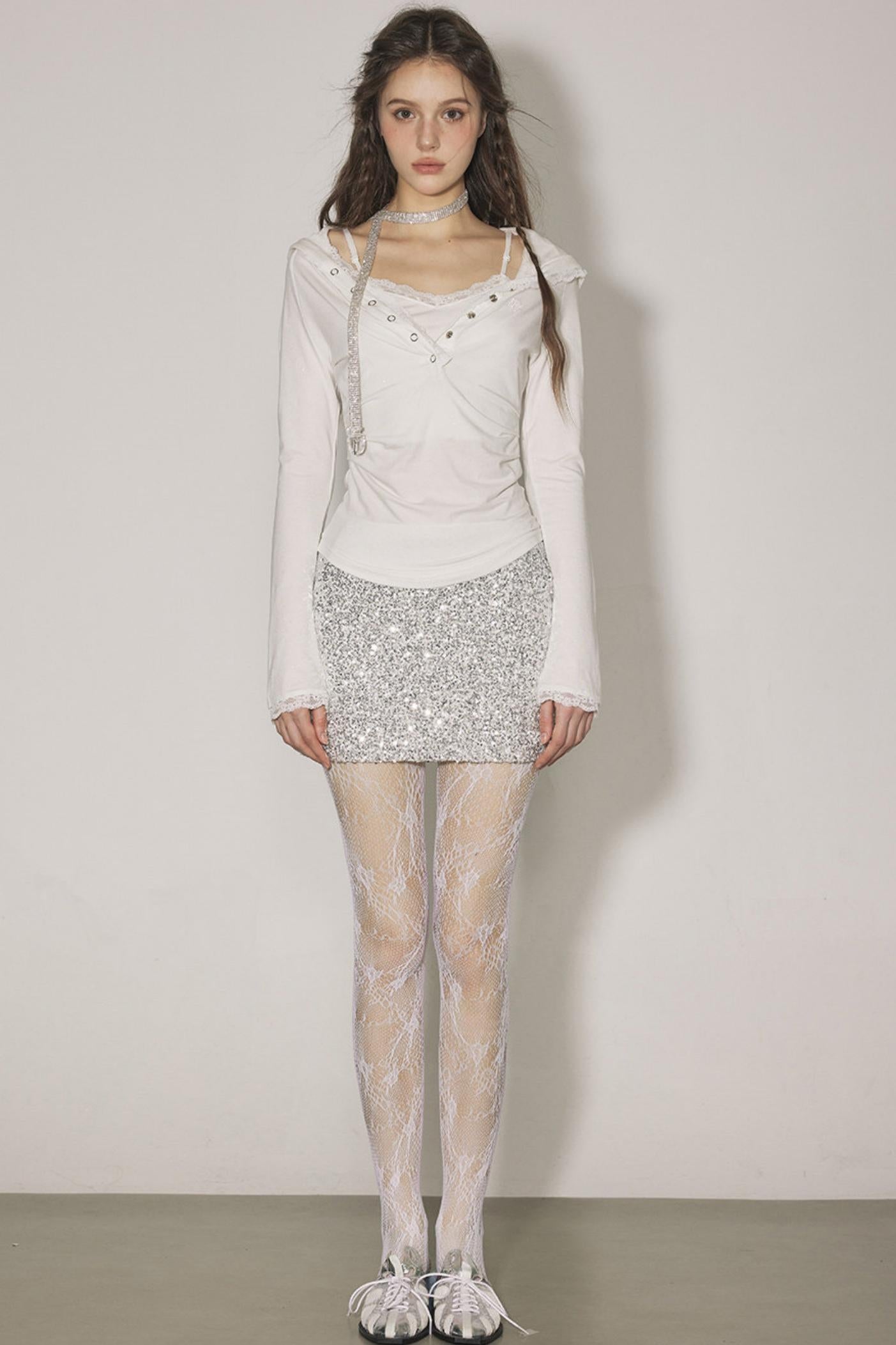 Woman wearing a white long-sleeve top, silver sequin skirt, and sheer stockings against a plain background