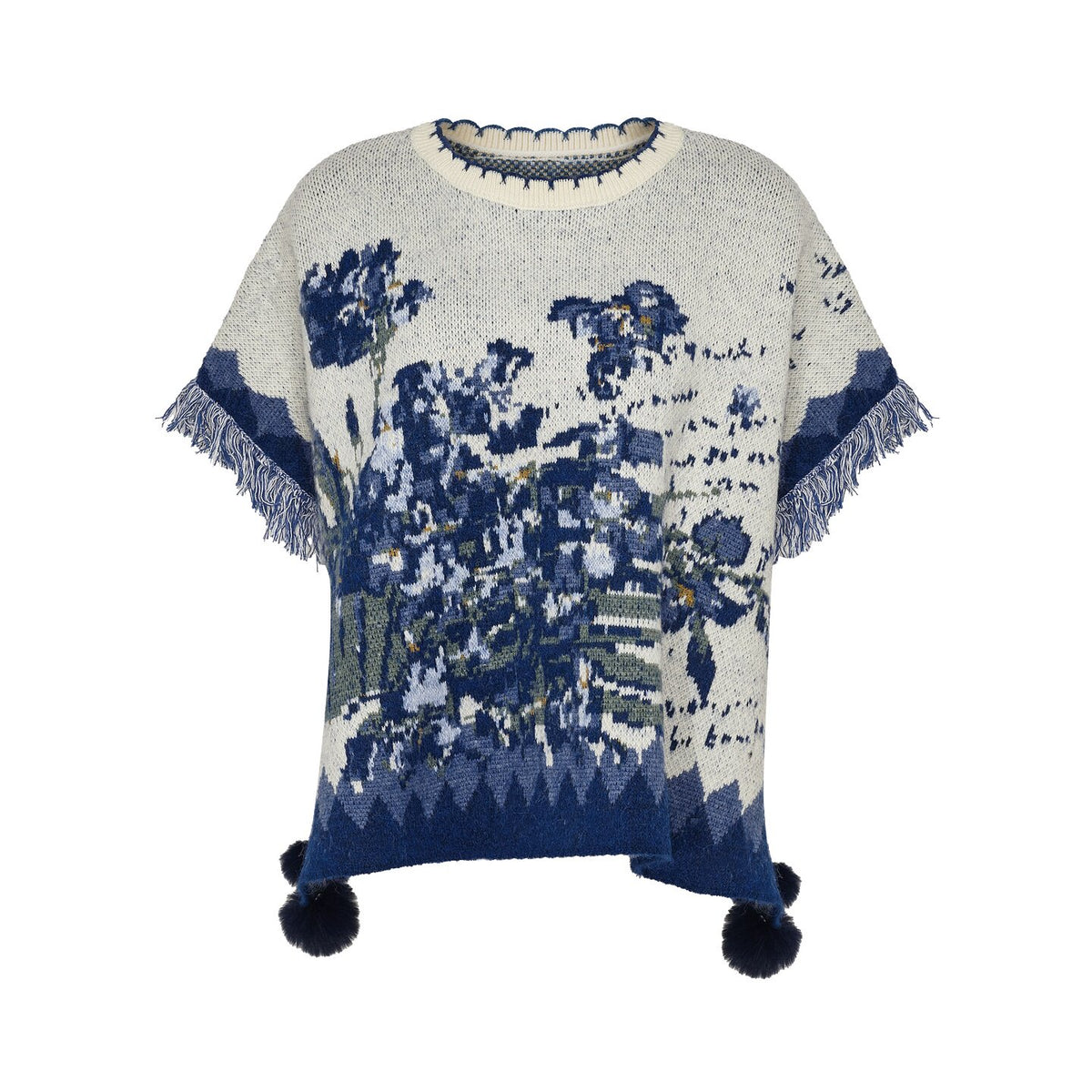 A cream-colored knit poncho sweater featuring a blue and navy blue botanical pattern, fringed edges, and dark pom-poms at the bottom, shown against a plain white background.
