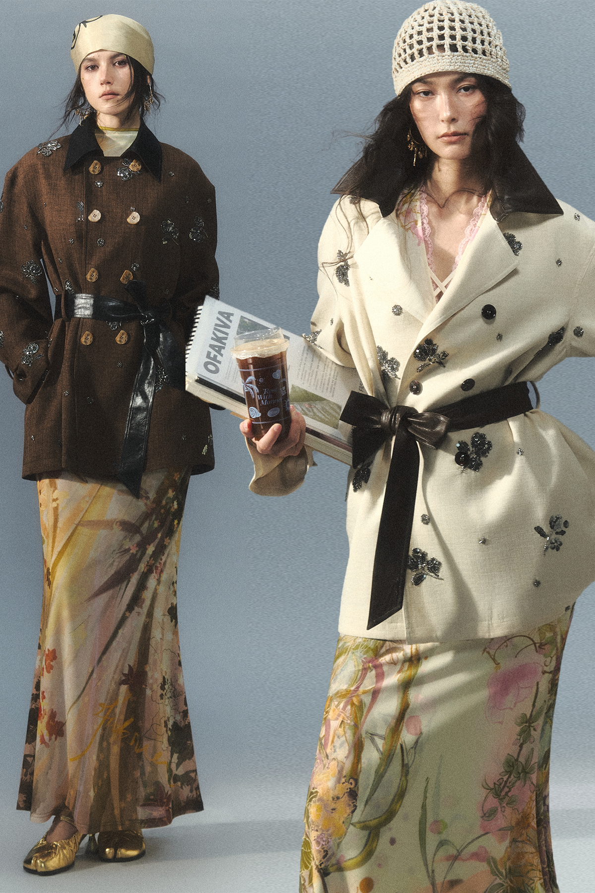 Two fashion models pose in editorial streetwear against a solid light gray studio background. The model on the left wears a brown double-breasted coat with a black collar, a wide belt, and a long beige skirt with a botanical print.