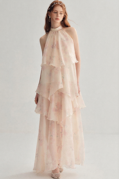 A woman modeling a sleeveless, high-neck, tiered maxi dress in a very light blush pink with a soft floral print, set against a plain light gray studio background.